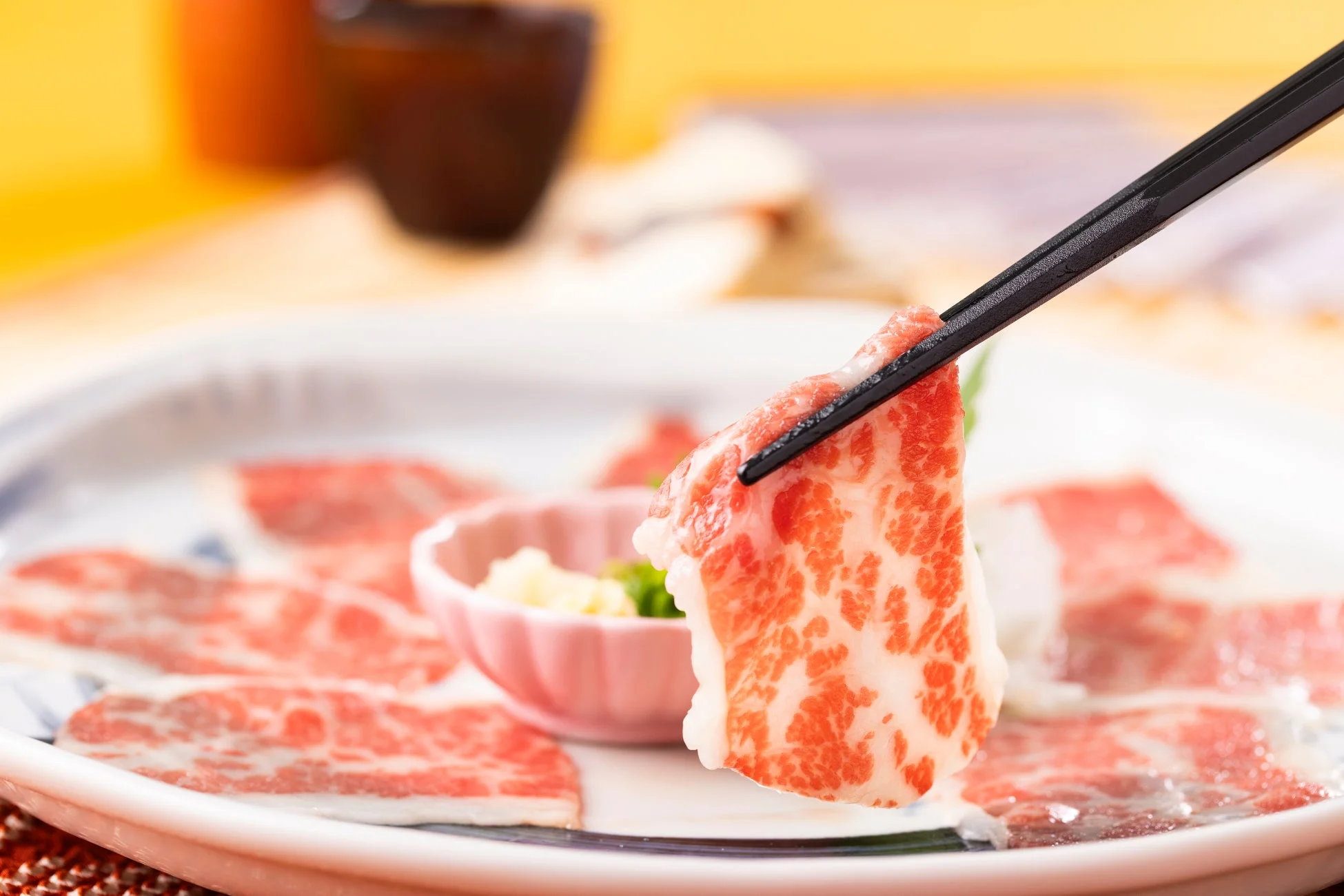 箸で持ち上げられた霜降りの生牛肉が主役の画像。皿には他にも薄切りの牛肉が並べられており、しゃぶしゃぶやすき焼きを連想させる。新鮮で上質な肉の質感が伝わる一枚。