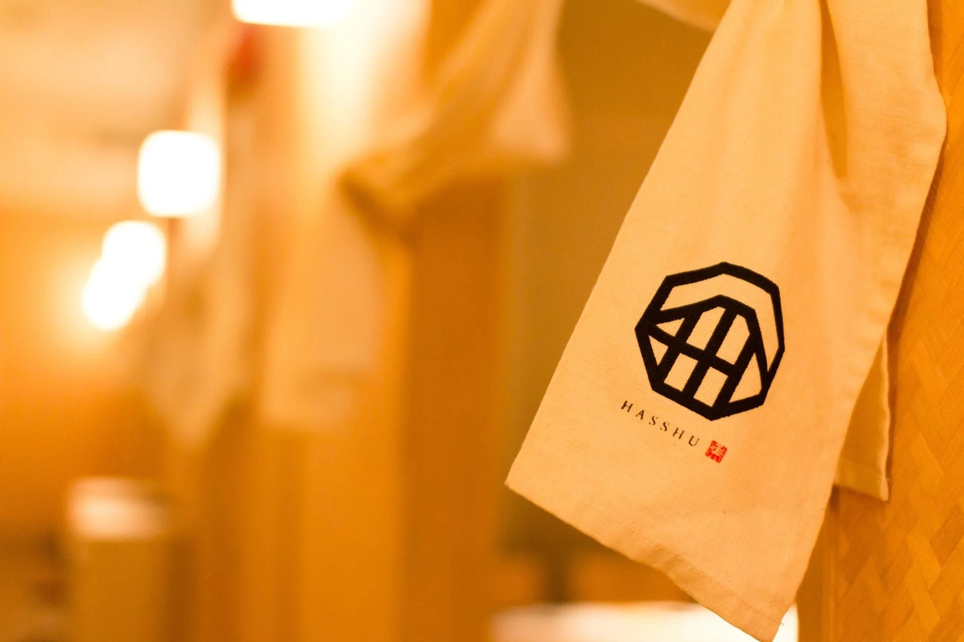 暖色系の照明がぼんやりと灯る空間で、幾何学的なロゴと「HASSHU」の文字がプリントされた薄い色の布が掛けられている様子を捉えた画像です。