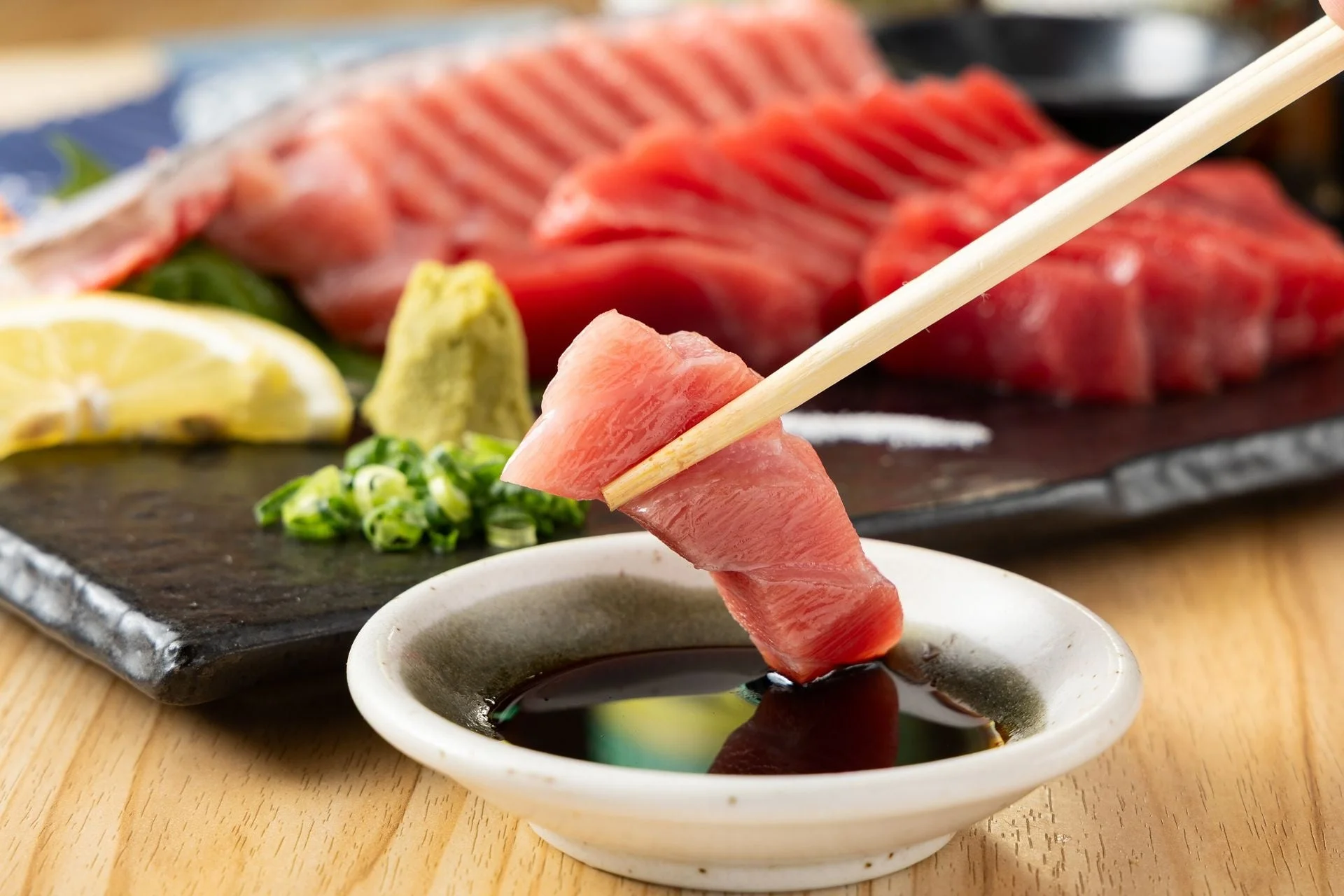 箸で新鮮なマグロの刺身を醤油につけている瞬間を捉えた画像。背景にはさらに多くのマグロの切り身、ワサビ、レモン、刻みネギが美しく盛り付けられている。
