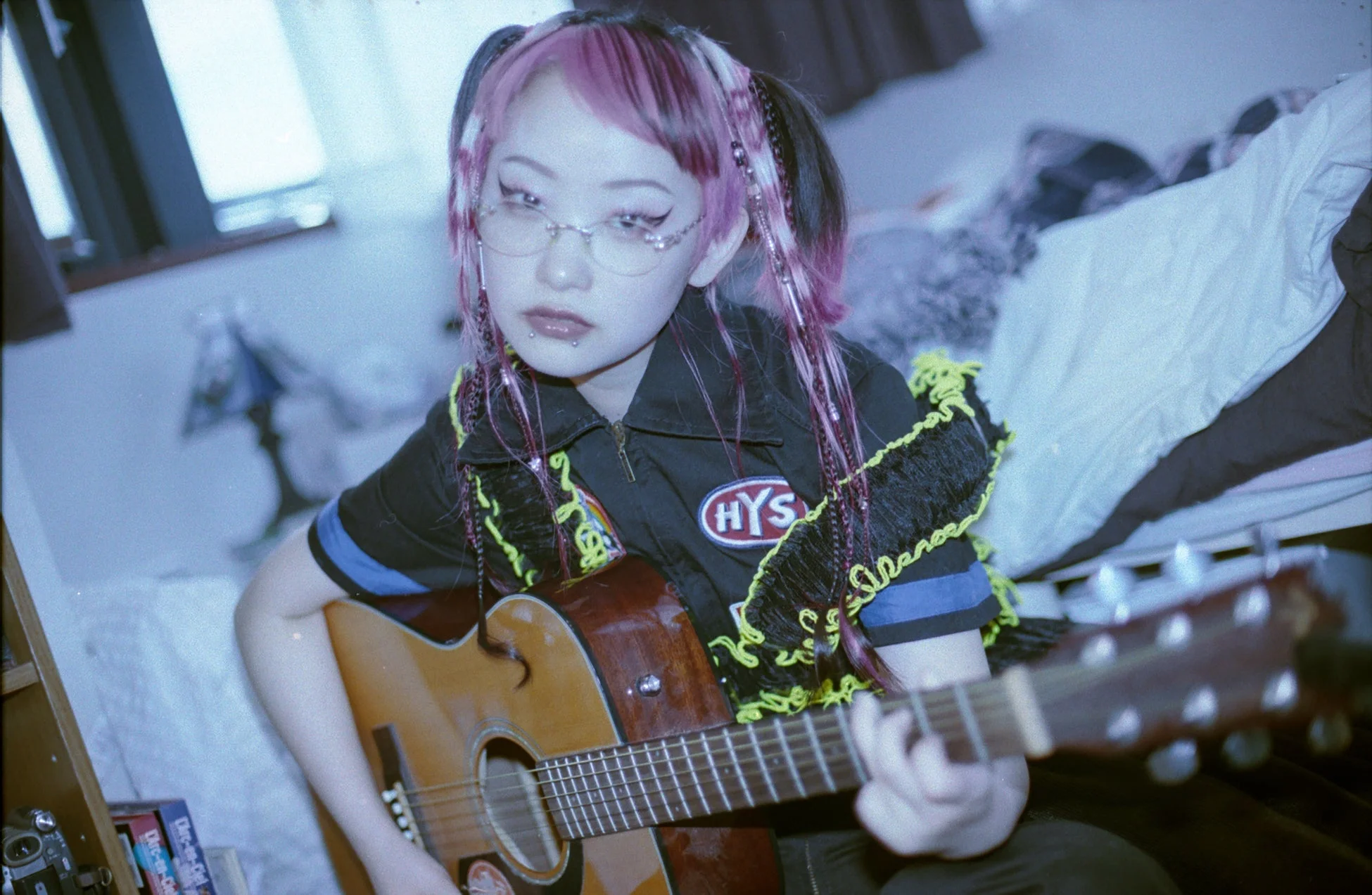 ピンクと黒のツインテールにメガネをかけた女性が、部屋でアコースティックギターを弾いています。