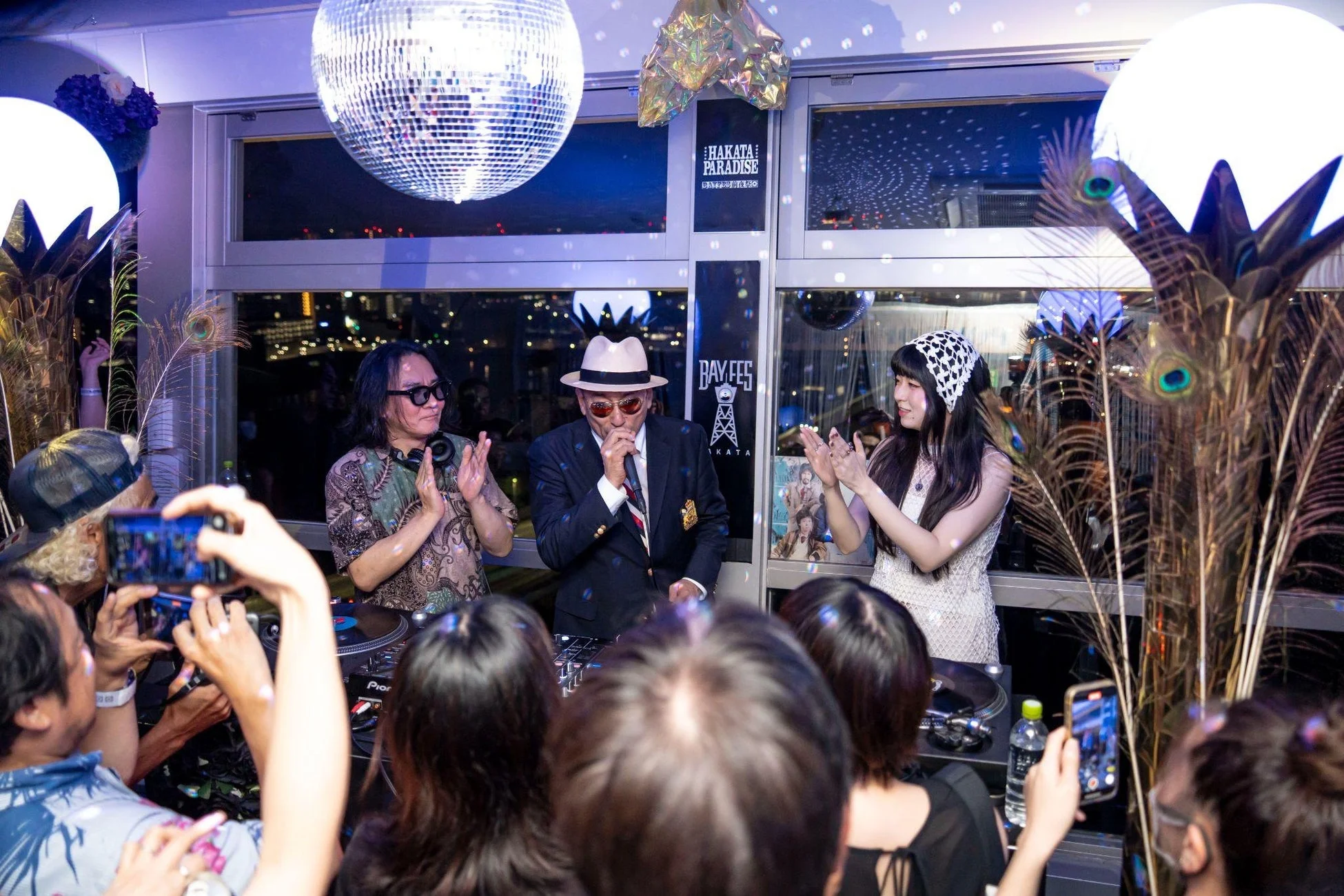ディスコボールが輝く会場で、DJブースを中心に人々が集まり、男性がマイクで話し、他の二人が拍手している様子