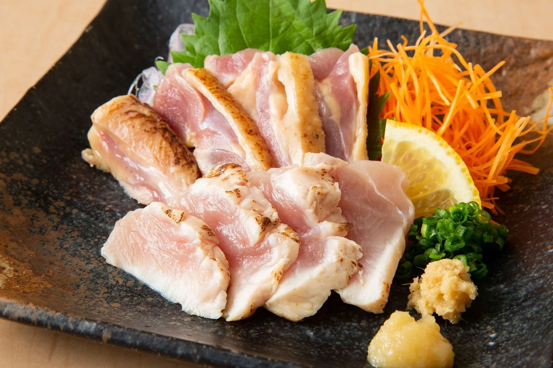 黒い皿に盛り付けられた新鮮な鶏のたたきです。皮目を炙った鶏もも肉と、白い鶏むね肉の刺身が美しく並べられています。添えられた大葉、千切り人参、レモン、小ねぎ、生姜、にんにくが彩りを添え、食欲をそそる一品です。