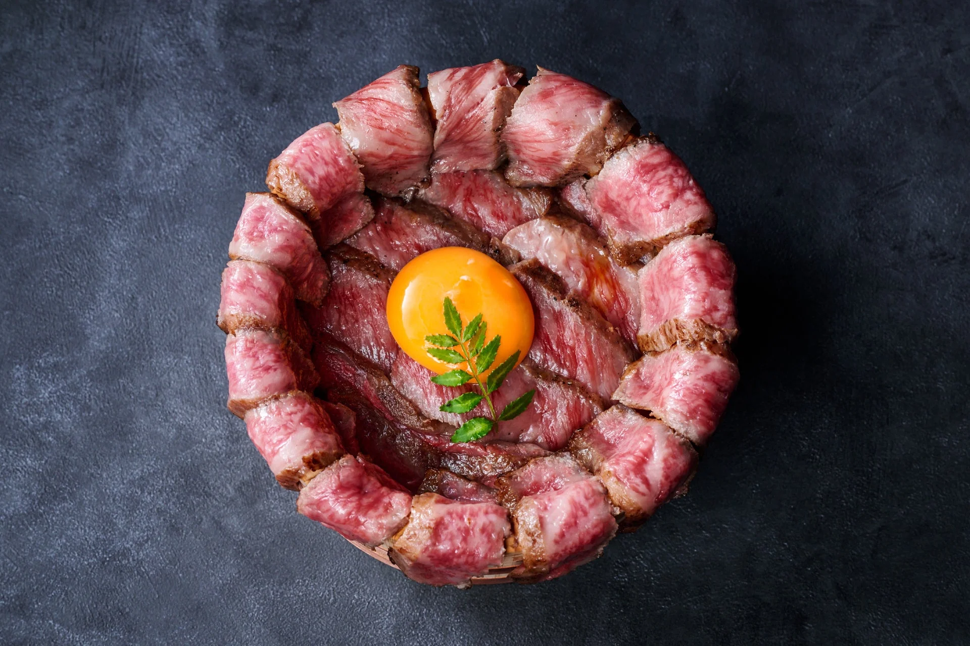 薄切りの牛肉が美しく盛り付けられた丼で、中央には鮮やかな卵黄と緑の葉が添えられています