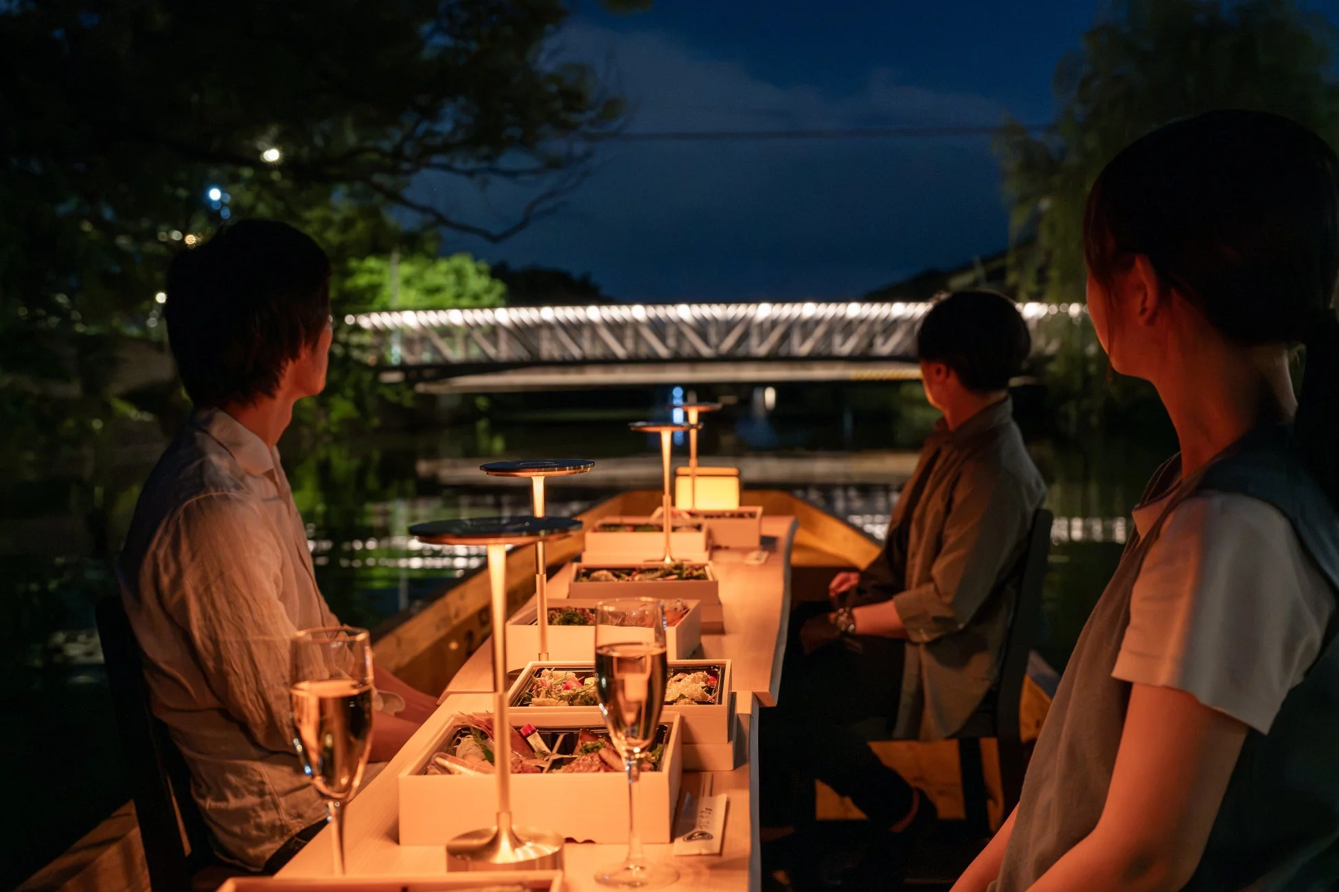 夜の水辺で、ライトアップされた橋を背景に、ボートの上で食事を楽しむ人々の様子。お洒落な料理とシャンパングラスが並び、ロマンチックなディナータイムが演出されている。