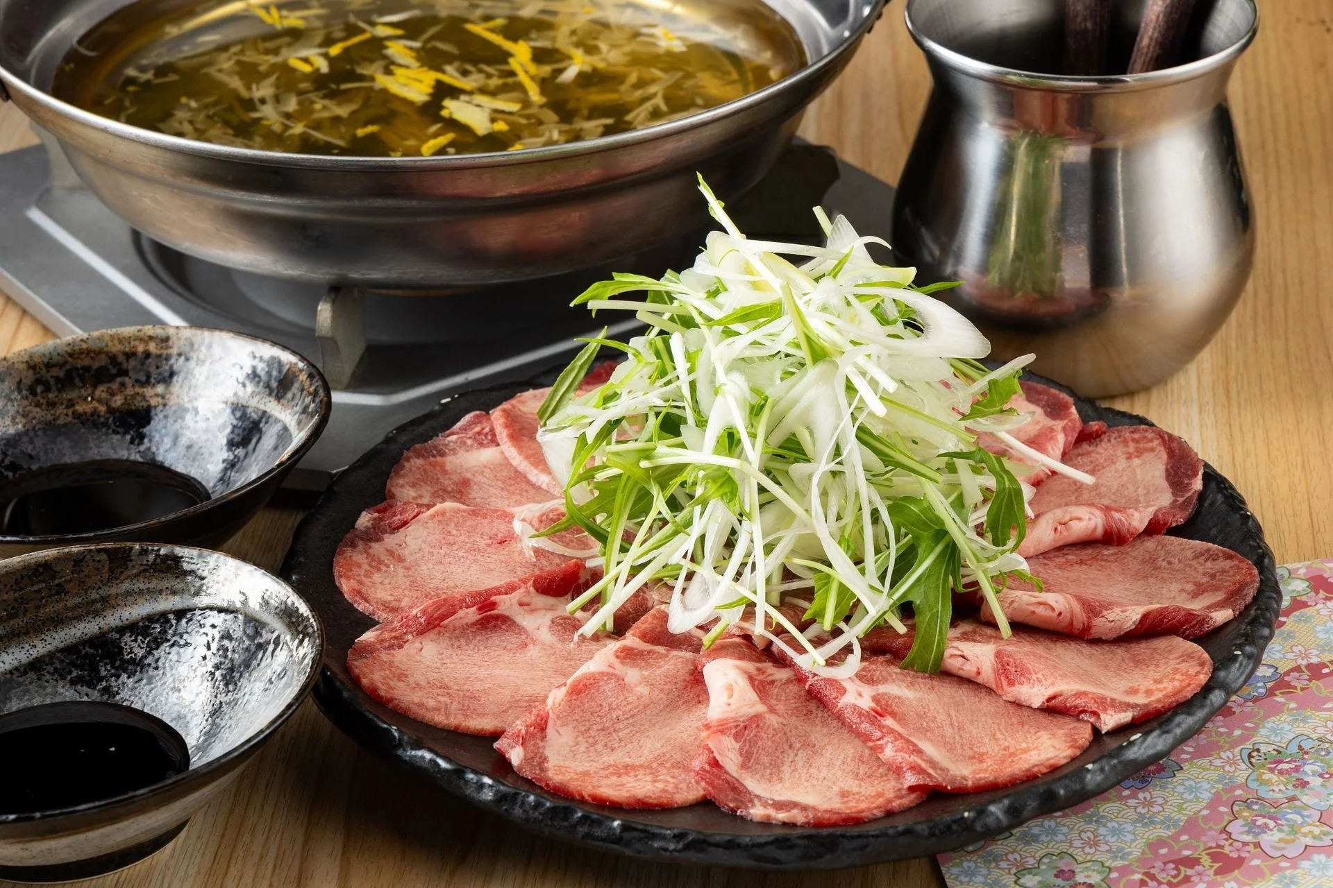 テーブルの上に置かれた鍋料理のセットです。薄切りの肉が盛り付けられた皿には、たっぷりの刻みネギと水菜が添えられています。奥には熱そうな出汁が入った鍋と、手前にはタレが入った小鉢が二つあります。