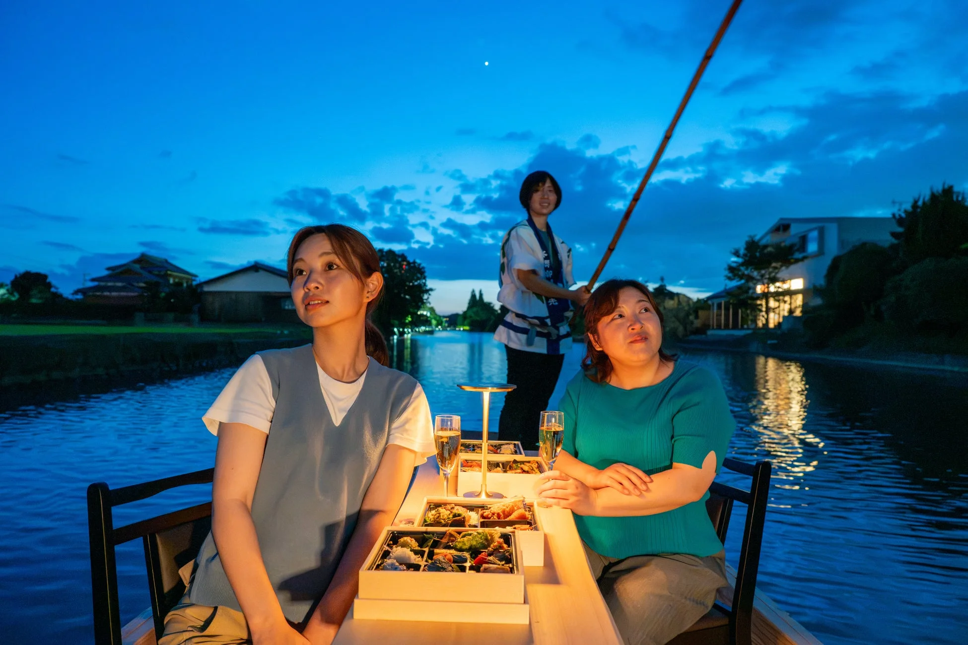 夕暮れ時の運河で、二人の女性が小舟に乗り、食事と飲み物を楽しんでいます。船頭もおり、水辺の風景を眺めながらのんびりとした時間を過ごしている様子が伺えます。