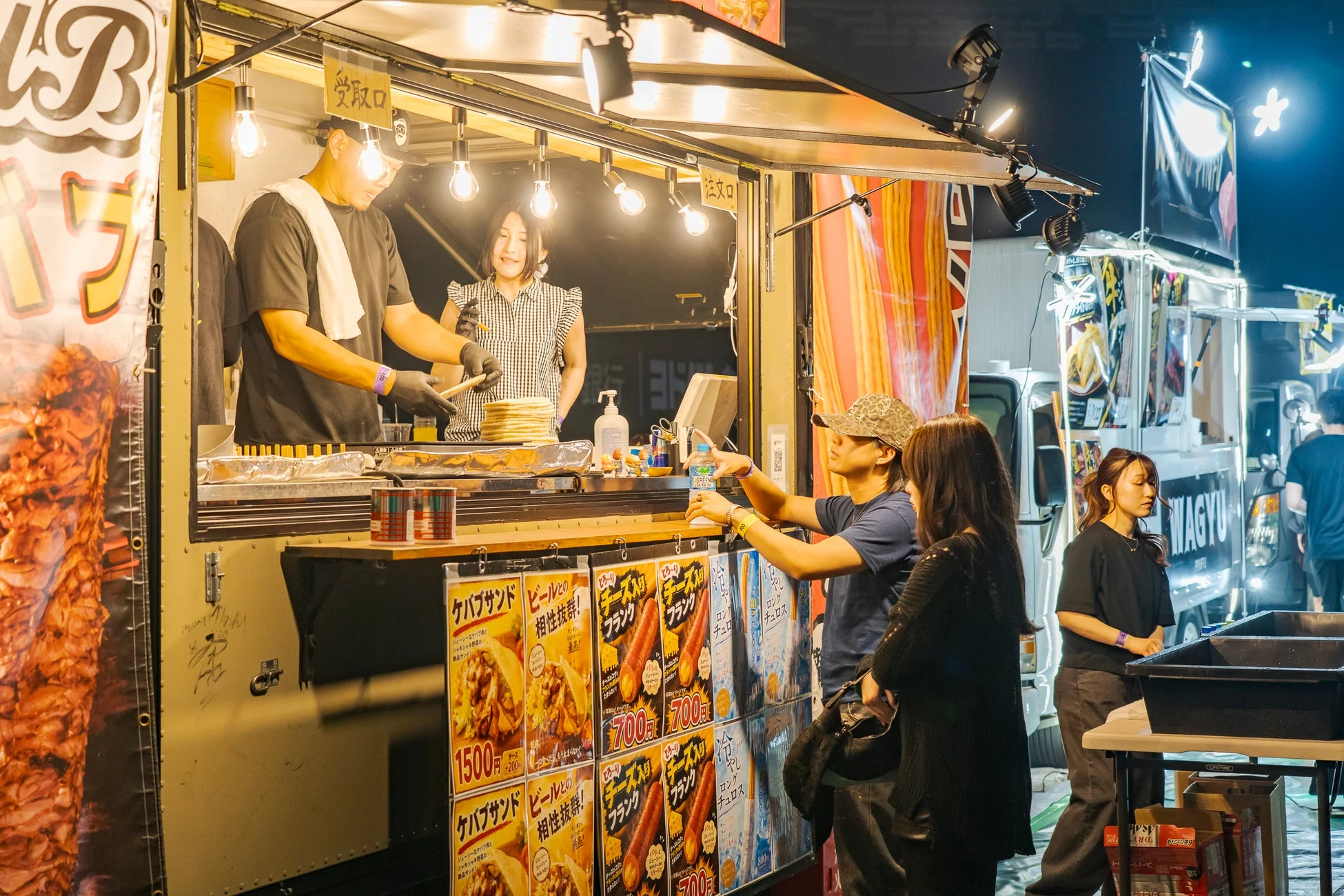 夜の屋外イベントで賑わうケバブのフードトラックの様子。店員が料理を提供し、客が注文や商品を受け取っている。ケバブサンドやチーズフランクなどのメニューが並び、活気ある雰囲気が伝わる。