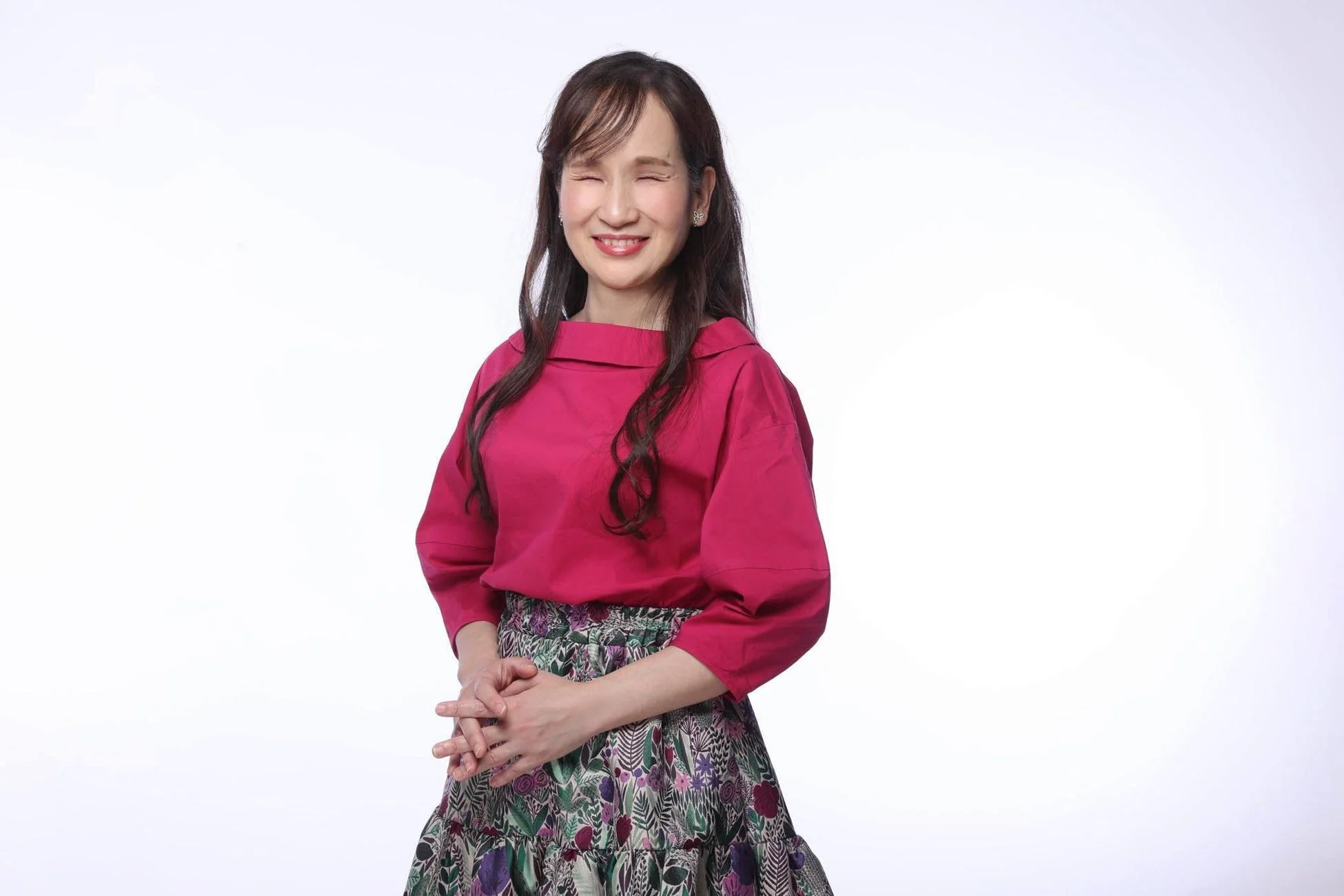 白い背景の前で、目を閉じてにこやかに微笑む女性の全身像です。