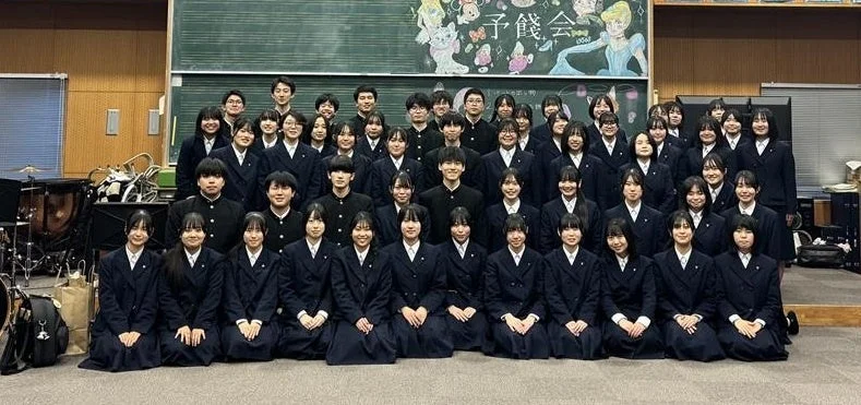 制服を着た大勢の学生が音楽室のような場所で集合写真を撮っている。