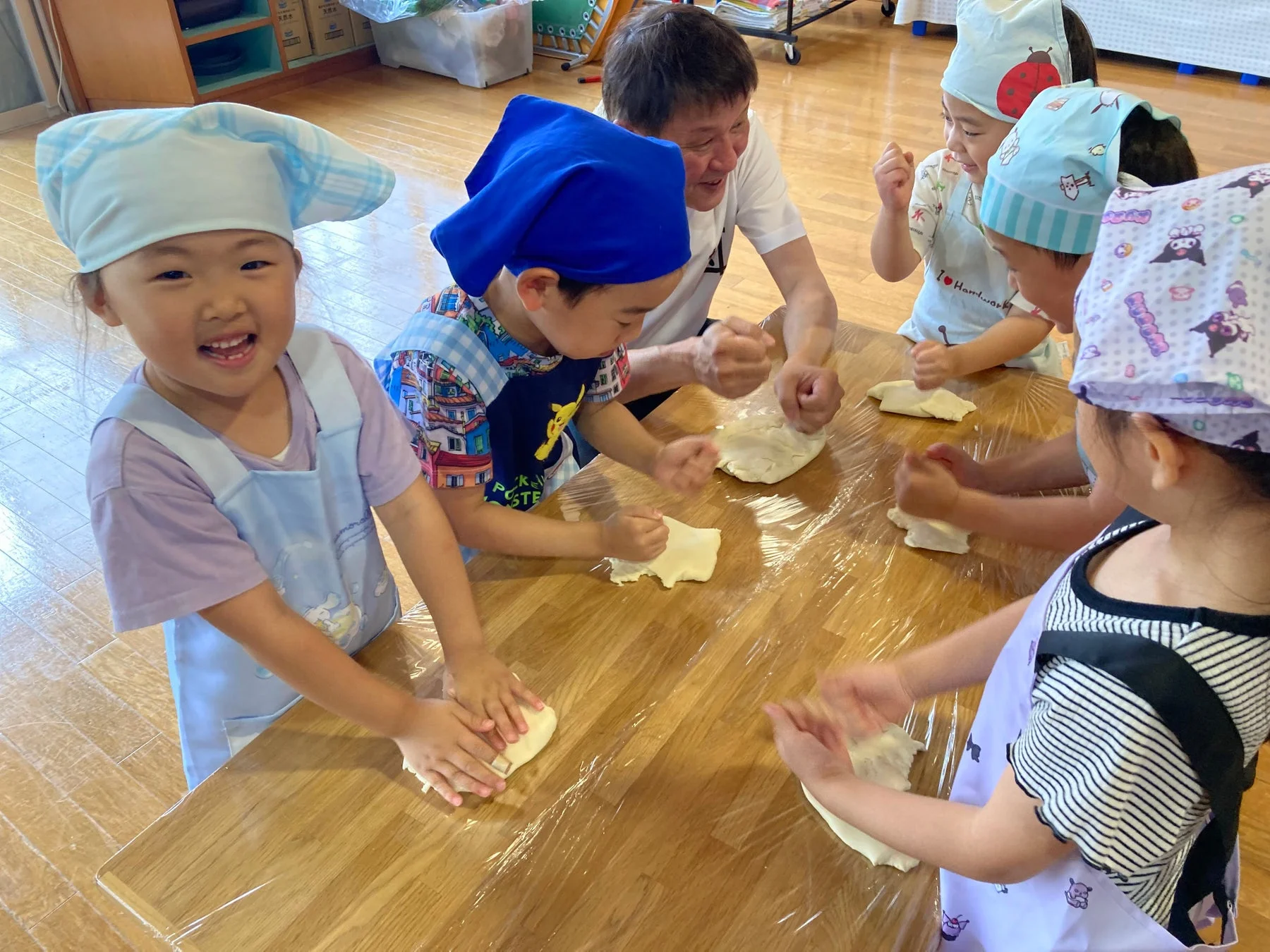 子供たちがエプロンとバンダナを身につけ、大人の指導のもと、テーブルで生地をこねるクッキング体験をしている様子。