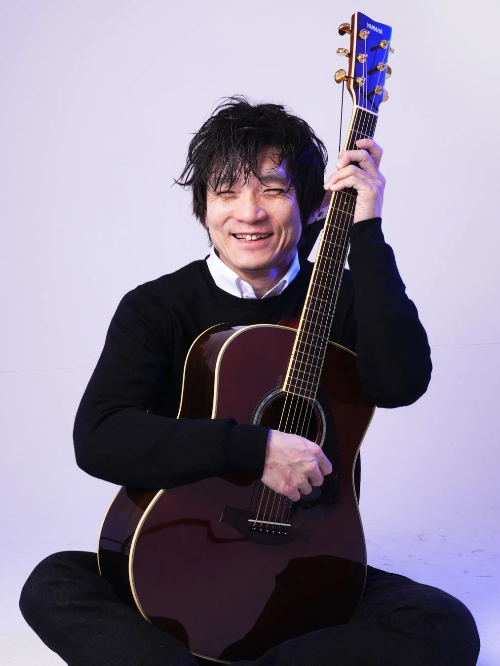 アコースティックギターを抱え、目を閉じて満面の笑みを浮かべる男性のポートレート。
