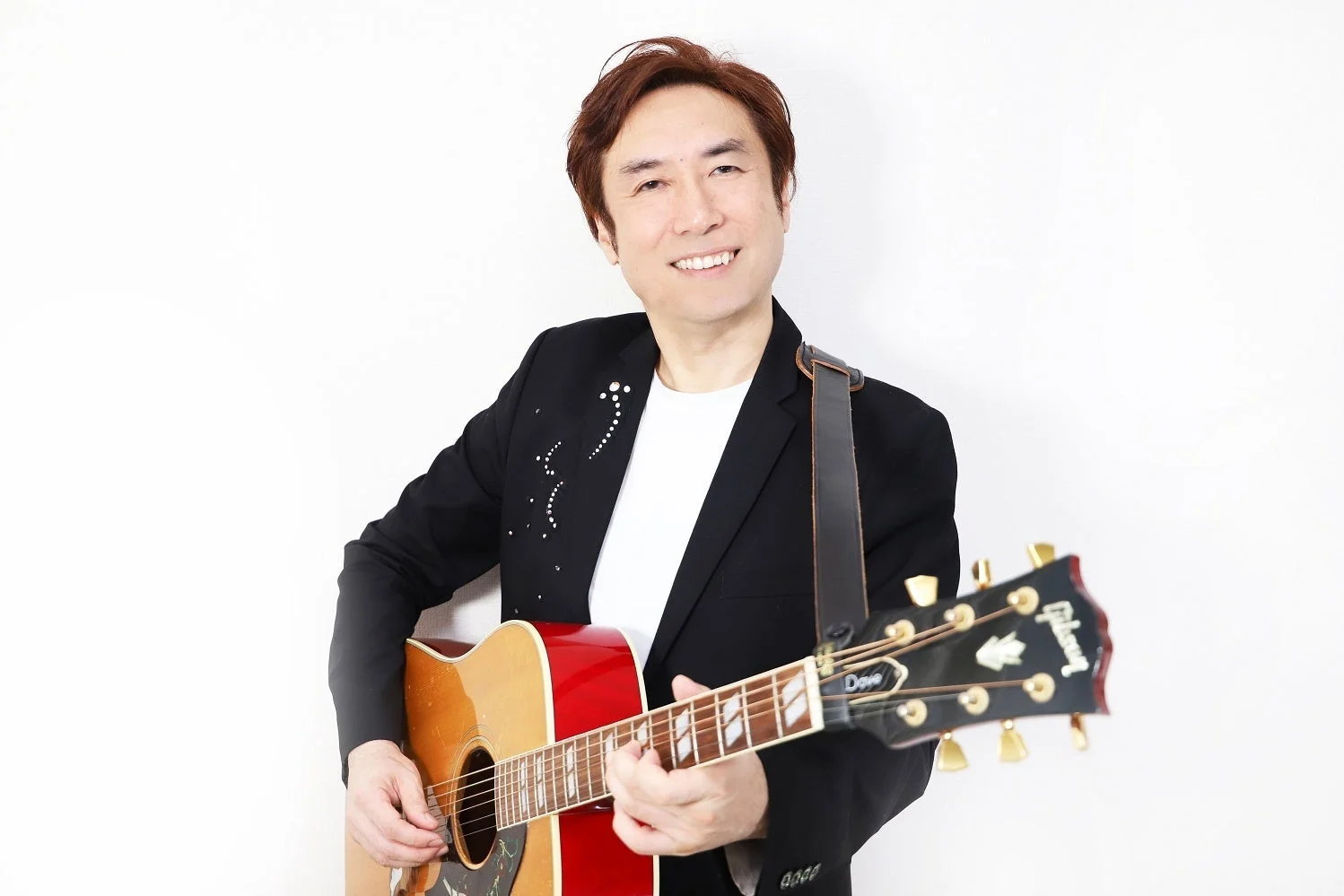 白い背景の前でアコースティックギターを抱え、笑顔を見せる男性ミュージシャンのポートレート写真です。