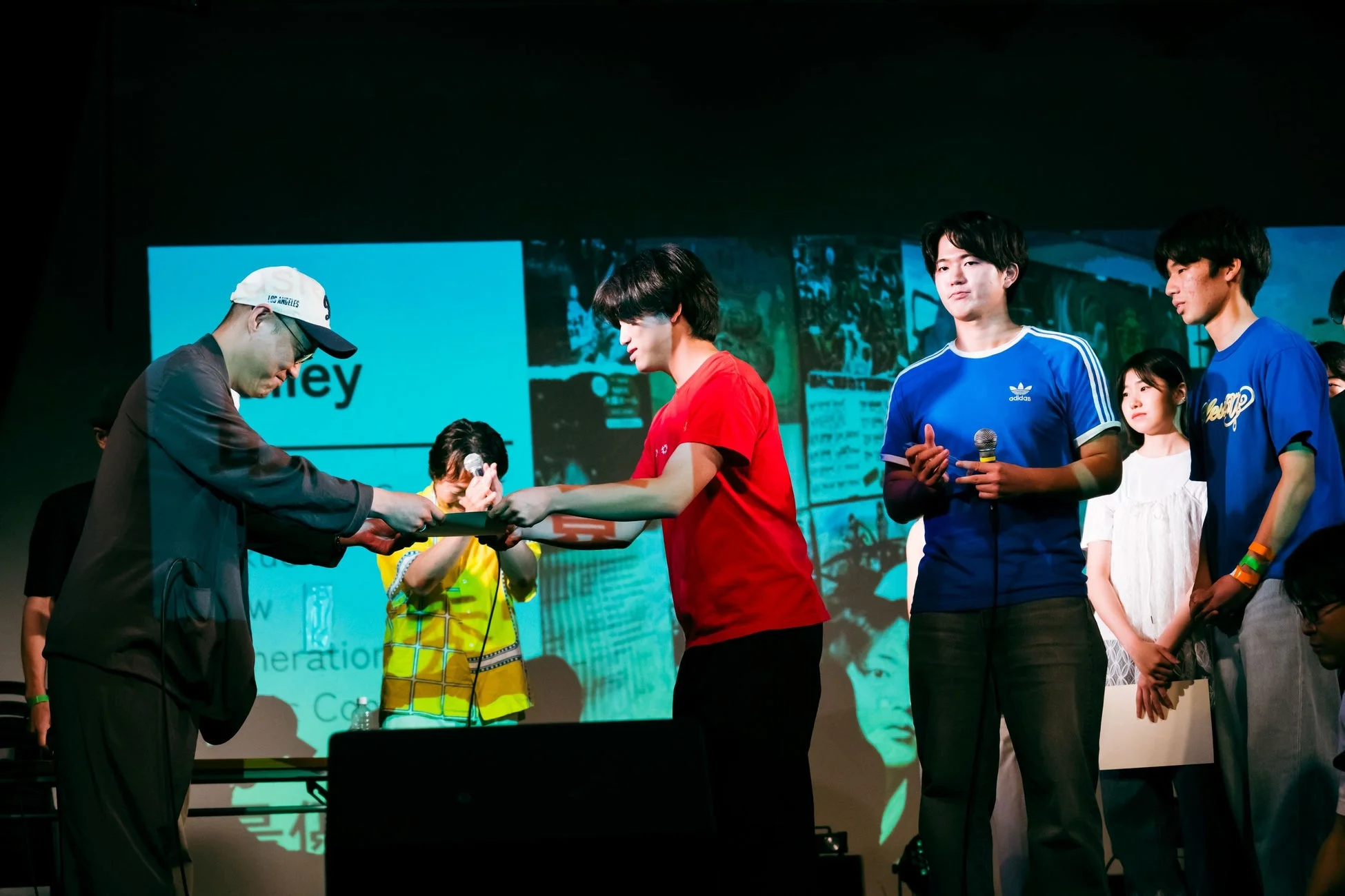 ステージ上で行われているイベントの一幕で、キャップをかぶった男性が若者に何かを手渡している様子。