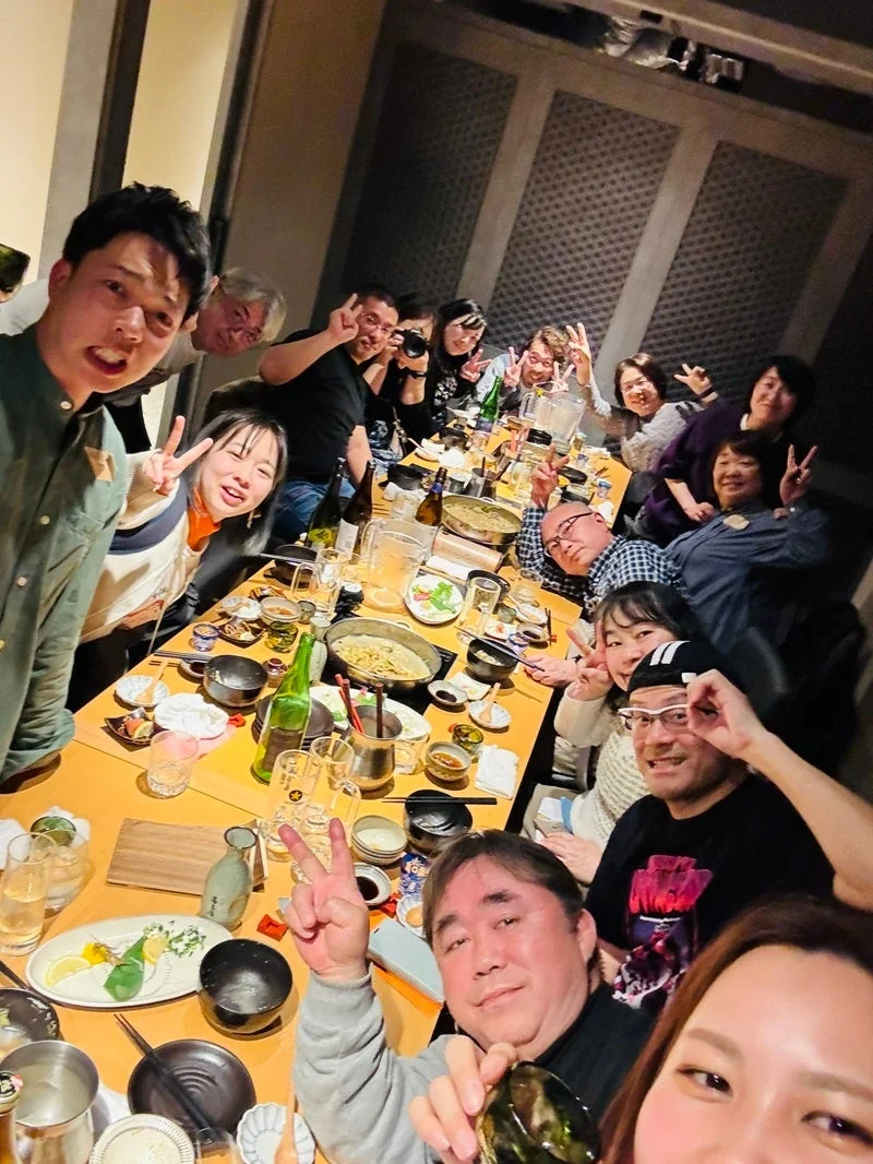居酒屋のような場所で、多くの人々がテーブルを囲んで食事や飲み物を楽しんでいる様子。皆笑顔でピースサインをしており、和やかな雰囲気の集合写真です。