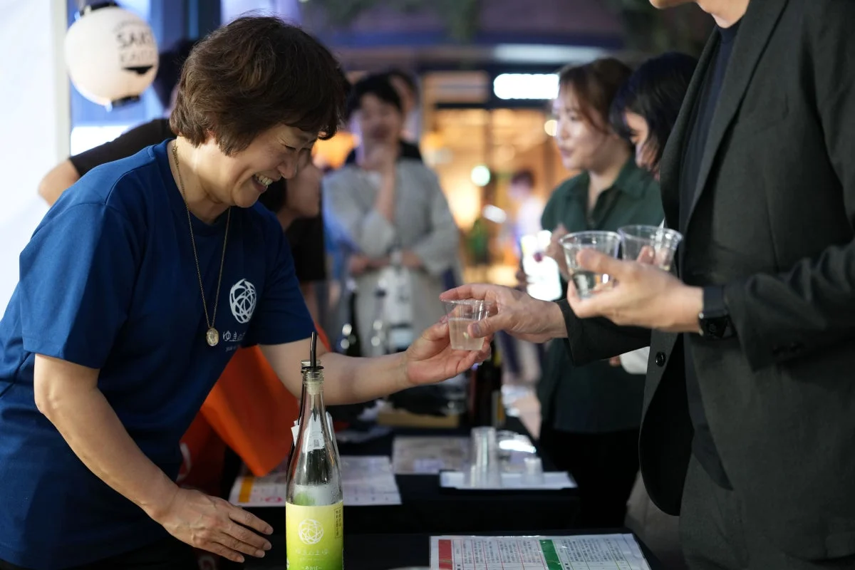 日本酒の試飲イベントで、笑顔の女性が参加者に日本酒を注いでいる様子です。青いTシャツを着た女性が緑色のボトルから酒を小さなカップに提供しており、会場は賑やかな雰囲気です。