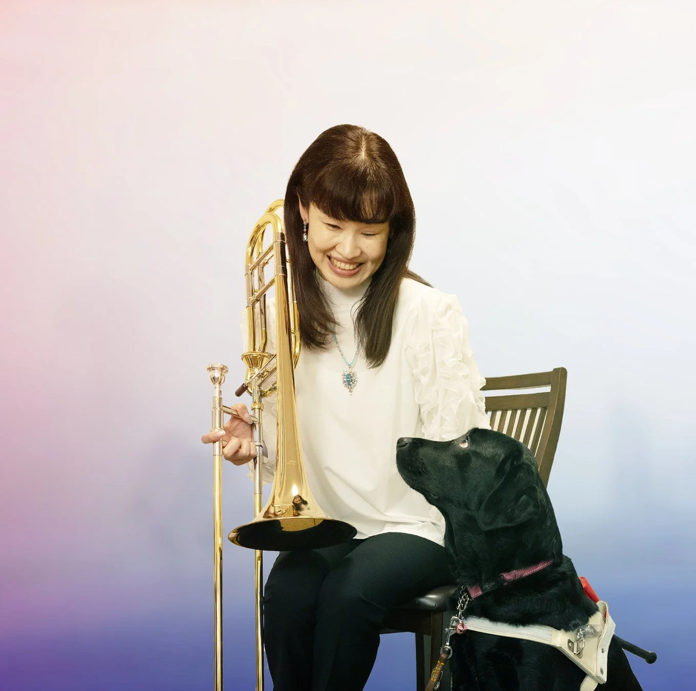 トロンボーンを持つ笑顔の女性と、彼女を見上げる黒い盲導犬が写っています。