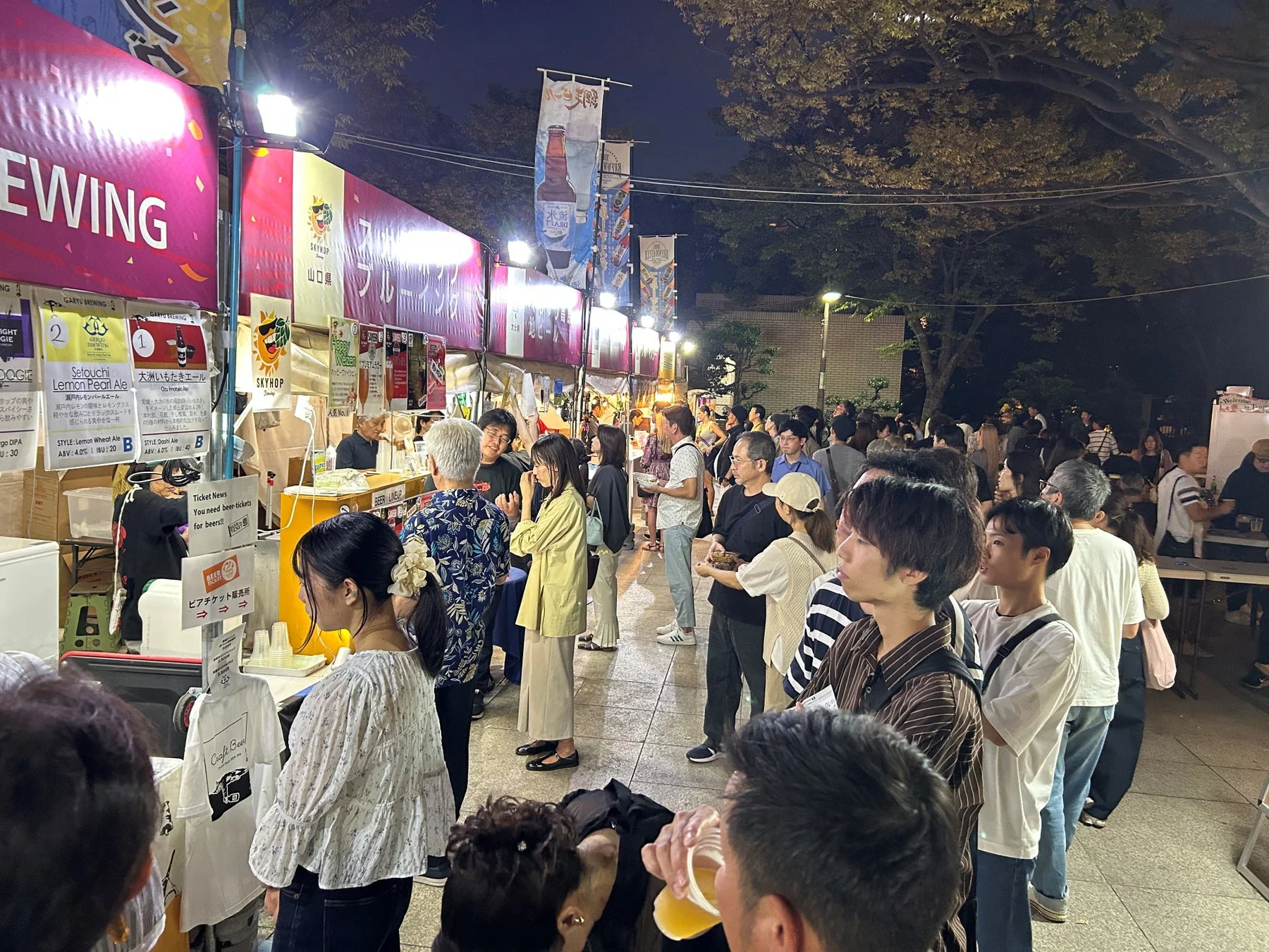 夜の屋外イベントで、多くの人々がクラフトビールの屋台に並んでいます。