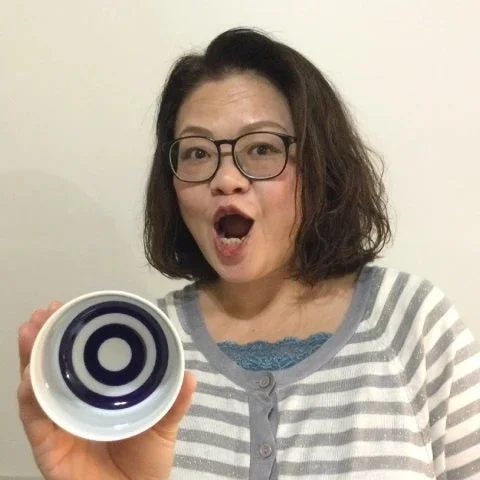 メガネをかけた中年女性が、口を大きく開けて驚いたような表情で、利き猪口を手に持っている画像です。白とグレーのストライプのカーディガンを着用しており、室内で撮影されたポートレートです。