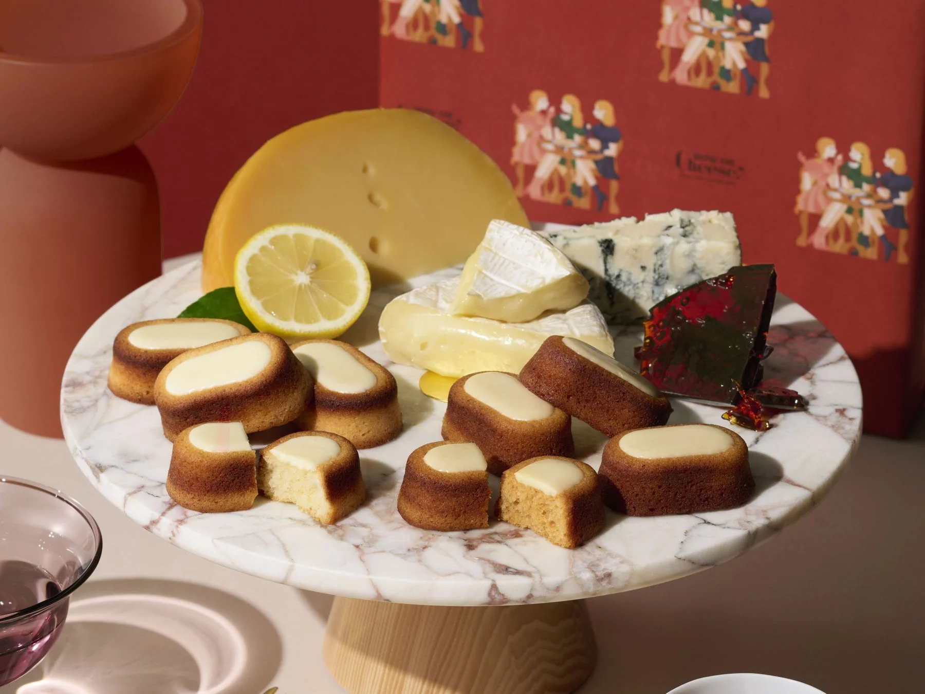 大理石のケーキスタンドに、数種類のチーズ、レモンのスライス、白いフロスティングの焼き菓子が盛り付けられています。赤を基調とした背景には人物のイラストが描かれています。