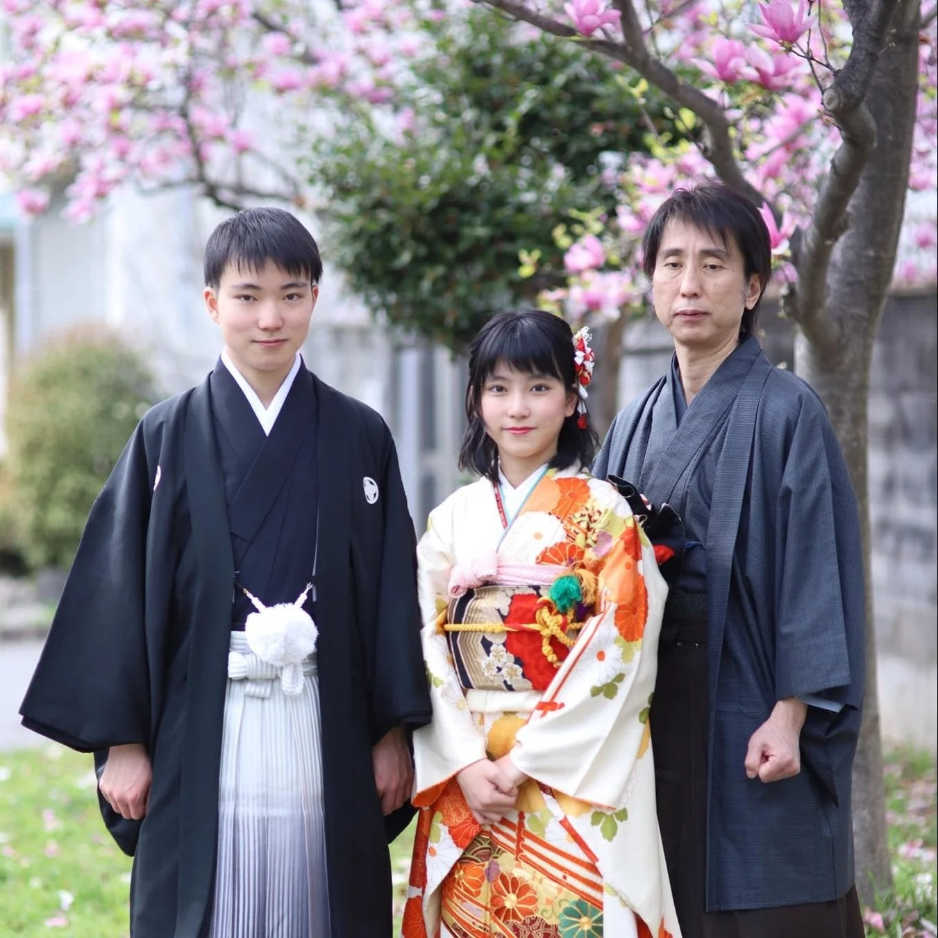 満開のピンク色の花が咲く木の下で、日本の伝統的な和装を身につけた家族3人が記念撮影をしている様子です。