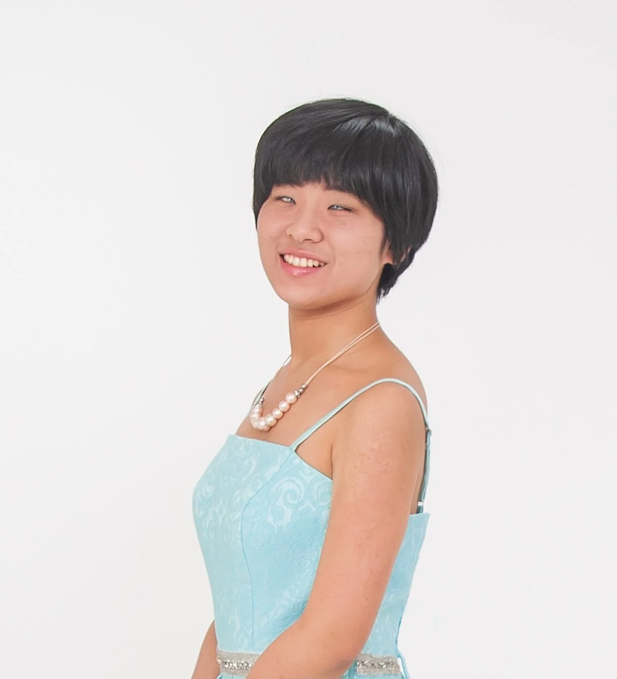 ショートヘアのアジア人女性が水色のドレスと真珠のネックレスを身につけ、白い背景の前で笑顔を見せています。