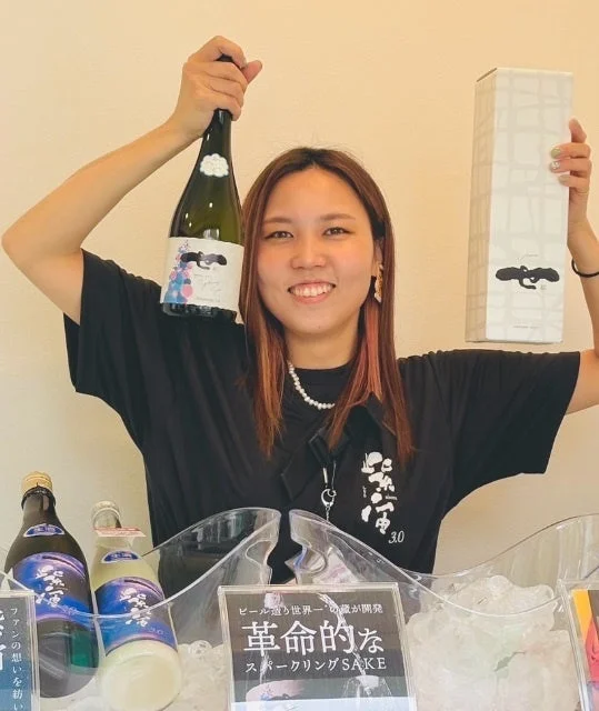笑顔の女性がスパークリング日本酒のボトルと箱を掲げています。手前には氷が入ったバケツに複数のボトルが並べられ、「革命的なスパークリングSAKE」と書かれたPOPが見えます。商品のプロモーションをしている様子です。