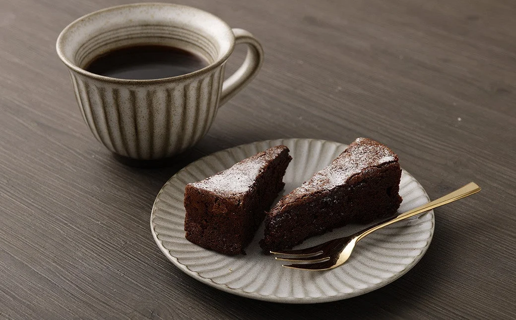 木製のテーブルに置かれたコーヒーカップと、粉砂糖がかけられたガトーショコラが2切れ盛り付けられた皿が写っています。温かい飲み物と甘いデザートでくつろぐ時間を連想させる一枚です。