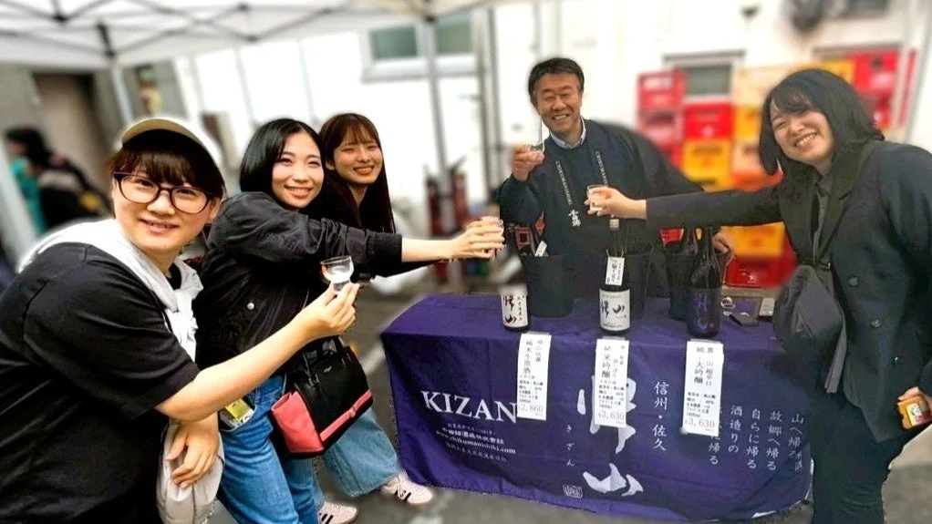 信州佐久のKIZAN酒造のブースで、人々が笑顔で日本酒の試飲を楽しむ屋外イベントの様子