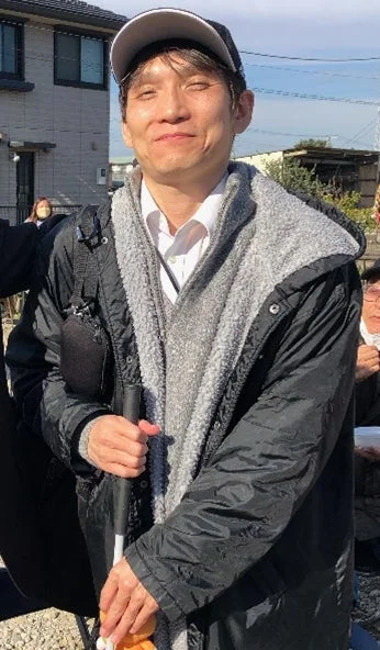 白い杖を持った男性が屋外で笑顔を見せている写真です。