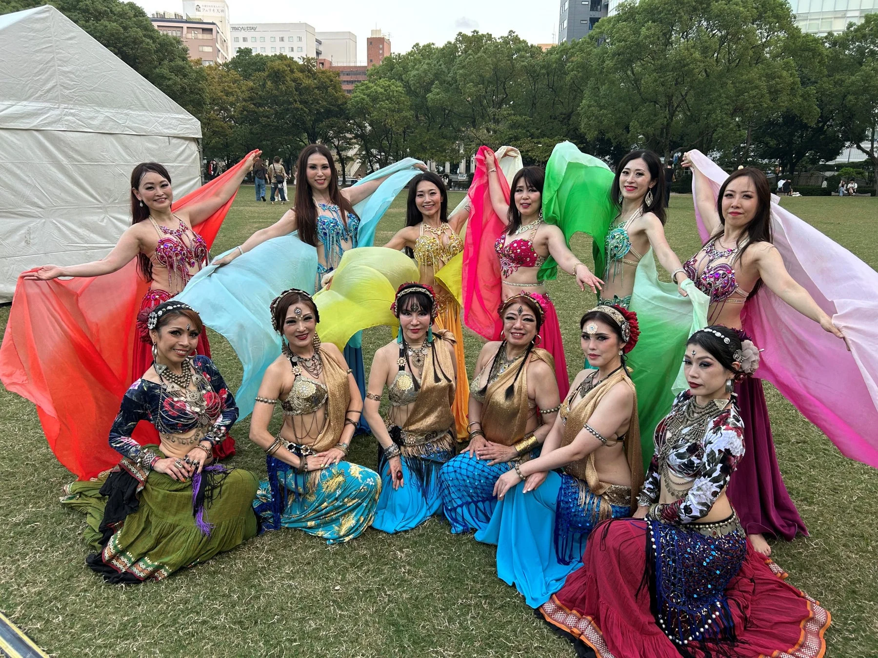屋外の芝生で、色鮮やかなベリーダンス衣装をまとった女性たちが、それぞれのスカーフを広げてポーズをとっています。