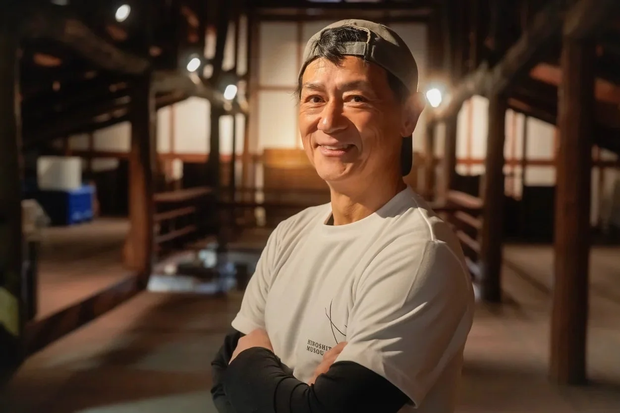 笑顔で腕を組んだ中年の男性が、木造の伝統的な建物の内部で立っているポートレート。親しみやすい表情が印象的です。