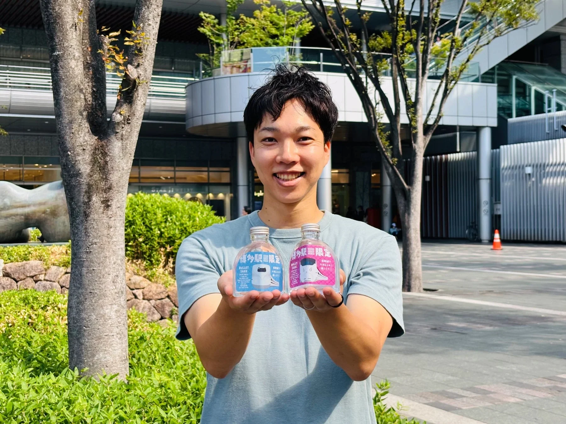 笑顔の男性が、新幹線デザインの「博多駅限定 BULLET SAKE」のボトルを2本手に持っている屋外の写真です。背景には現代的な建物と木々が写っています。