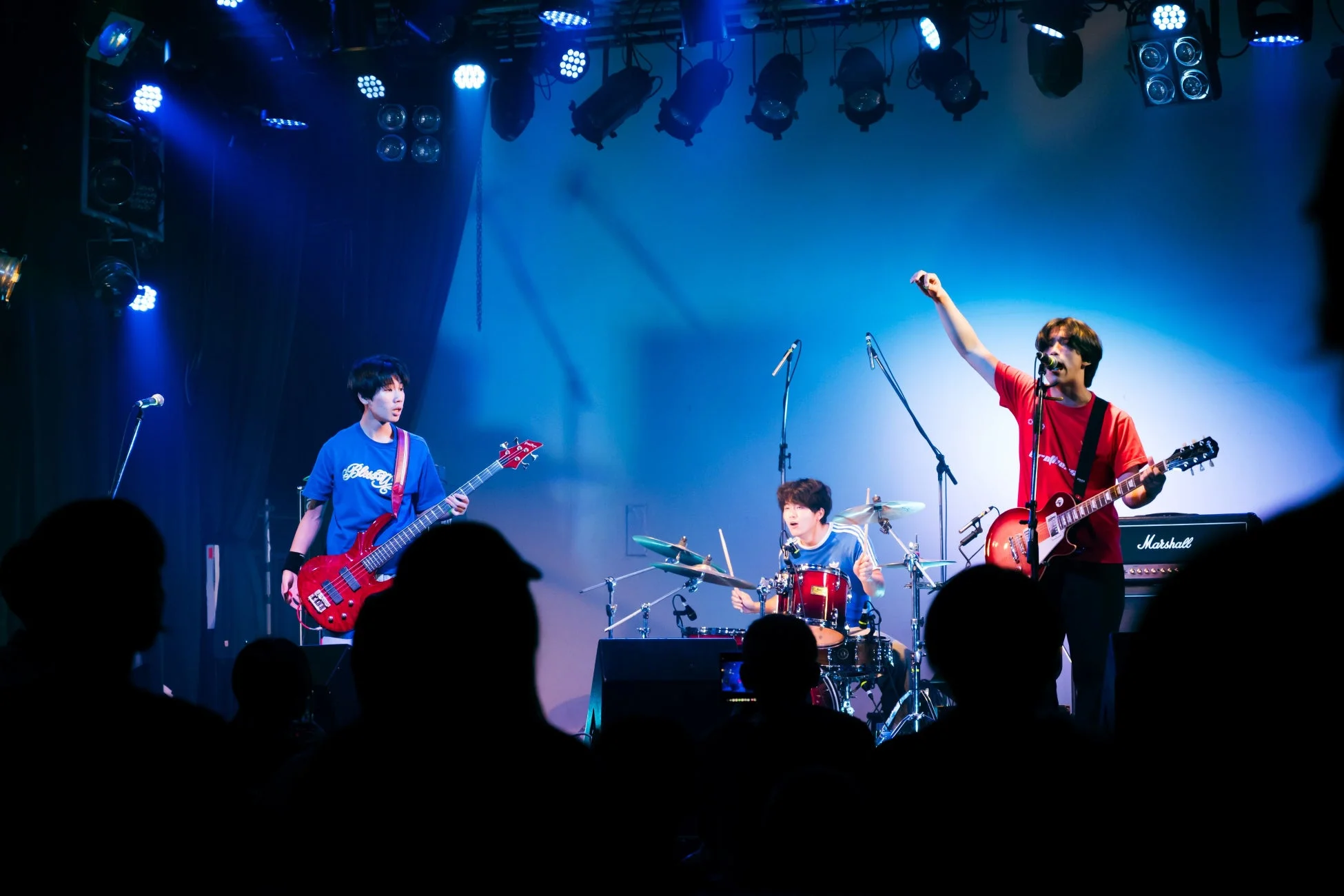 青い照明が照らすライブステージで、3人組のロックバンドが熱い演奏を披露しています。