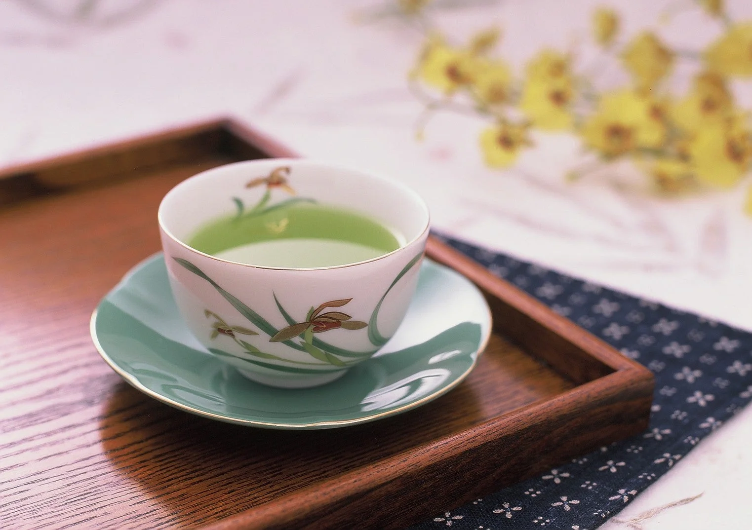 美しい花柄の湯呑みに注がれた緑茶が、木製のお盆と和柄の布の上に置かれています。背景のぼやけた黄色い花が、落ち着いた和の雰囲気を醸し出しています。