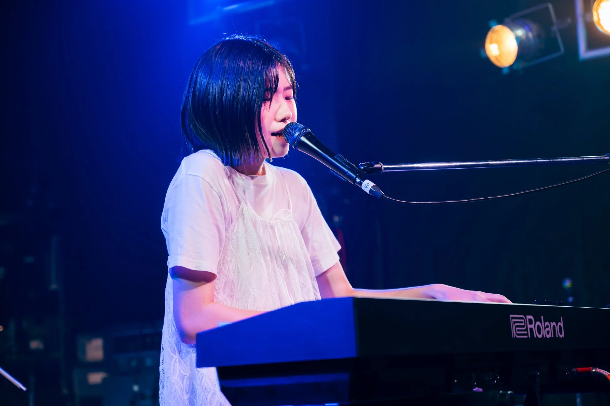 青い照明が印象的なステージで、白い服を着た女性がキーボードを演奏しながら歌っています。