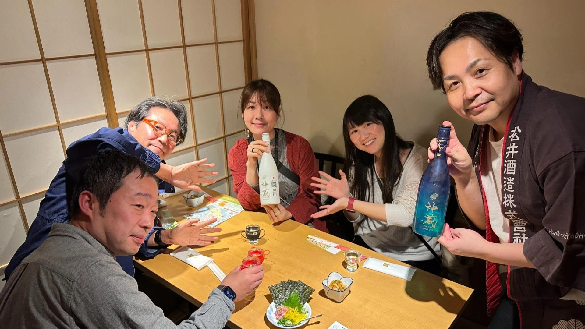 和風の個室で、男女5人がテーブルを囲んで食事や日本酒を楽しんでいる様子