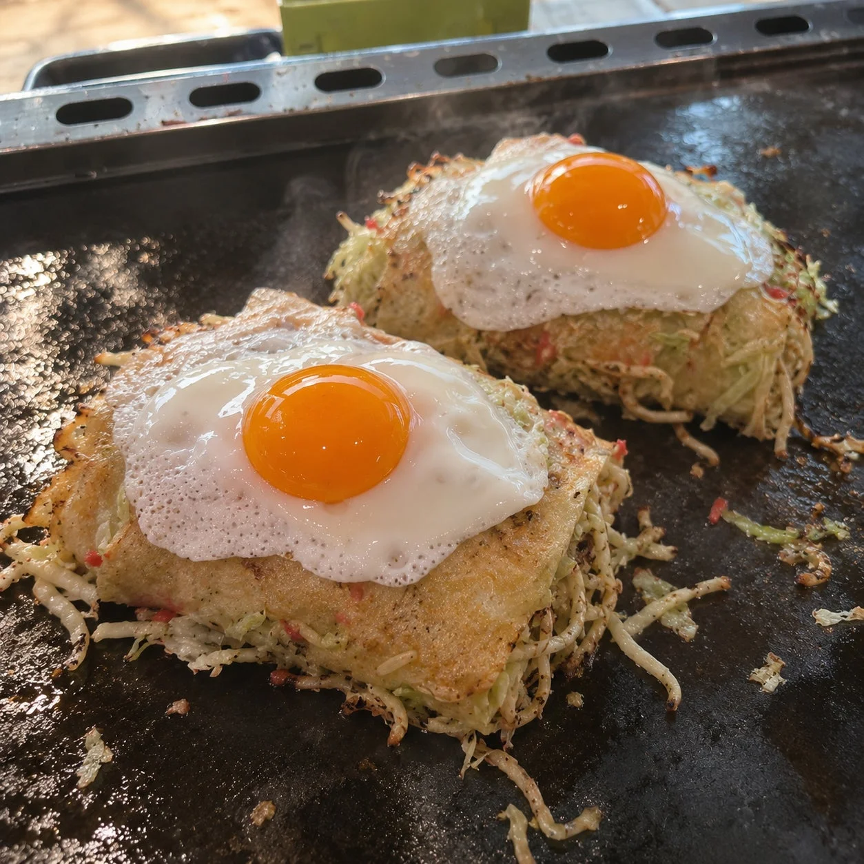 熱い鉄板の上で調理されている、目玉焼きが乗ったお好み焼きのような料理