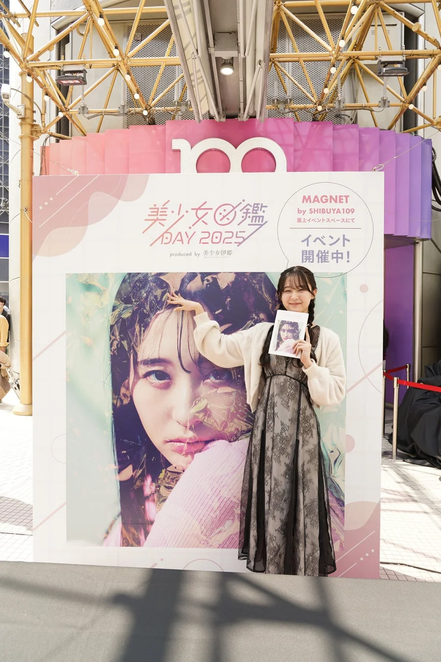 「美少女図鑑 DAY 2025」のイベント会場で、大きなポスターの前に立つ若い女性