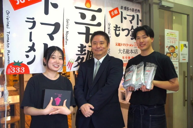 ラーメン店の前で、代表者とスタッフが笑顔で立つ写真