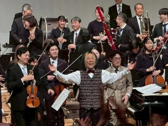 演奏を終えたオーケストラのメンバーがステージ上で笑顔を見せ、中央の人物が両手を広げて観客に感謝を伝えている様子。多くの楽器が確認でき、和やかな雰囲気が伝わる一枚です。